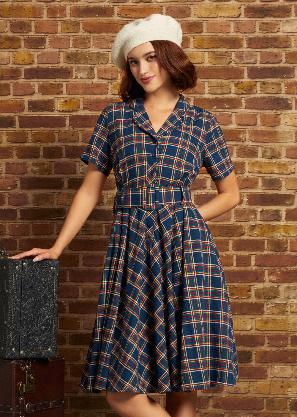 A woman wearing a vintage-style plaid dress and white beret stands against a brick wall, posing next to a vintage suitcase.