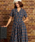 A woman wearing a vintage-style plaid dress and white beret stands against a brick wall, posing next to a vintage suitcase.
