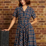 A woman wearing a vintage-style plaid dress and white beret stands against a brick wall, posing next to a vintage suitcase.