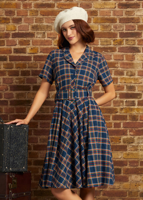 A woman wearing a vintage-style plaid dress and white beret stands against a brick wall, posing next to a vintage suitcase.