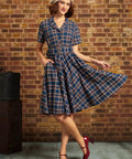 Woman wearing navy plaid cotton shirt dress with matching belt and side pockets, holding out the full skirt