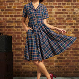 Woman wearing navy plaid cotton shirt dress with matching belt and side pockets, holding out the full skirt
