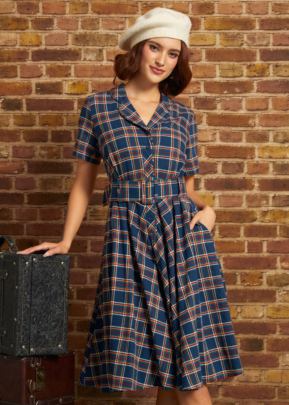 A woman wearing a blue plaid short-sleeve dress with a matching belt and a cream beret, posing next to a vintage suitcase against a brick wall background.