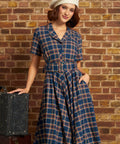 A woman wearing a blue plaid short-sleeve dress with a matching belt and a cream beret, posing next to a vintage suitcase against a brick wall background.