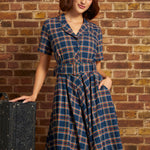 A woman wearing a blue plaid short-sleeve dress with a matching belt and a cream beret, posing next to a vintage suitcase against a brick wall background.