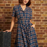 A woman wearing a blue plaid short-sleeve dress with a matching belt and a cream beret, posing next to a vintage suitcase against a brick wall background.