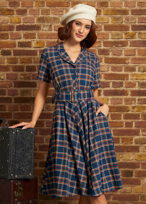 A woman wearing a blue plaid short-sleeve dress with a matching belt and a cream beret, posing next to a vintage suitcase against a brick wall background.