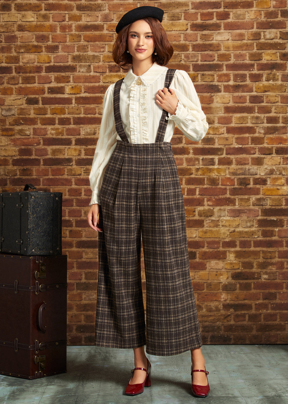A woman stands against a brick wall wearing a vintage-style outfit with a cream ruffled blouse, brown plaid suspenders, wide-leg cropped trousers, a black beret, and red Mary Jane heels. A vintage suitcase sits to her left.