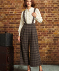 A woman stands against a brick wall wearing a vintage-style outfit with a cream ruffled blouse, brown plaid suspenders, wide-leg cropped trousers, a black beret, and red Mary Jane heels. A vintage suitcase sits to her left.