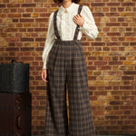 A woman stands against a brick wall wearing a vintage-style outfit with a cream ruffled blouse, brown plaid suspenders, wide-leg cropped trousers, a black beret, and red Mary Jane heels. A vintage suitcase sits to her left.