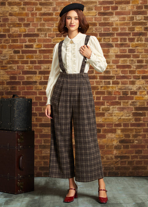 A woman stands against a brick wall wearing a vintage-style outfit with a cream ruffled blouse, brown plaid suspenders, wide-leg cropped trousers, a black beret, and red Mary Jane heels. A vintage suitcase sits to her left.