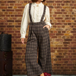 Woman wearing brown plaid wide-leg cropped suspender pants with an ivory ruffled blouse and red Mary Jane heels