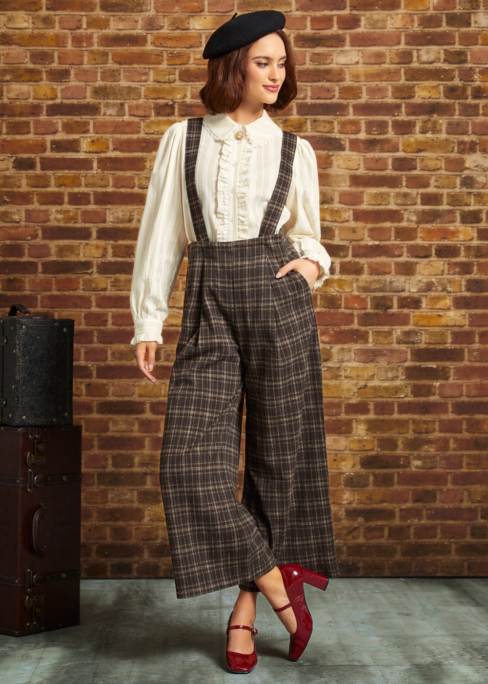 Woman wearing brown plaid wide-leg cropped suspender pants with an ivory ruffled blouse and red Mary Jane heels