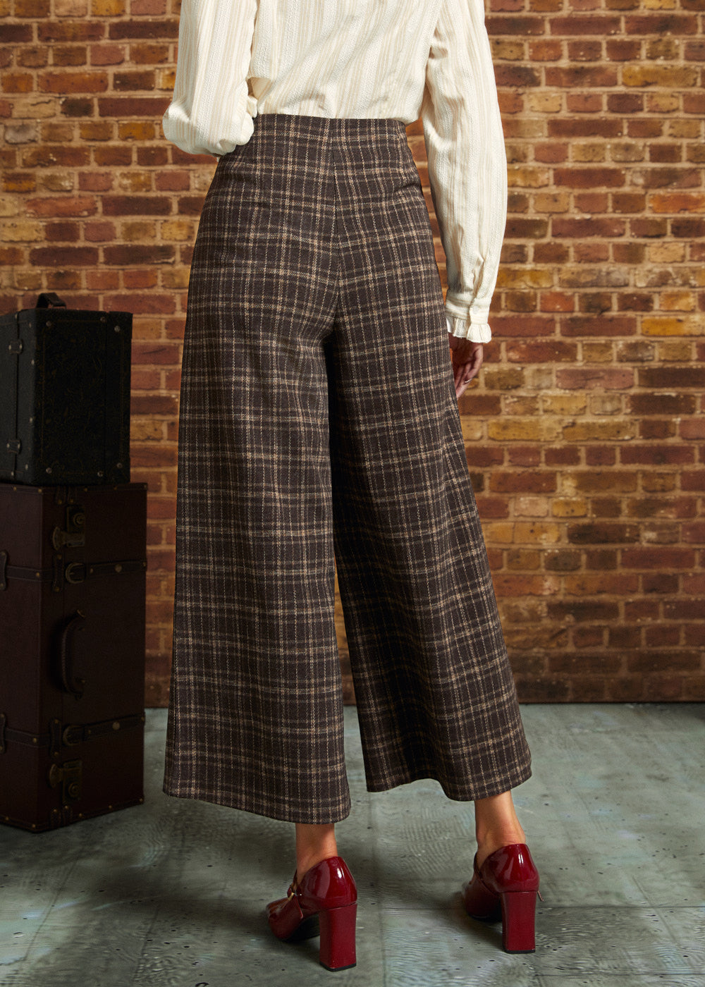 A person shown from the back wearing brown plaid cropped wide-leg trousers with a cream blouse and red heels, standing in front of a brick wall.