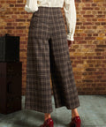 A person shown from the back wearing brown plaid cropped wide-leg trousers with a cream blouse and red heels, standing in front of a brick wall.