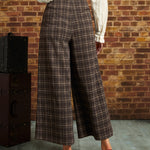 A person shown from the back wearing brown plaid cropped wide-leg trousers with a cream blouse and red heels, standing in front of a brick wall.