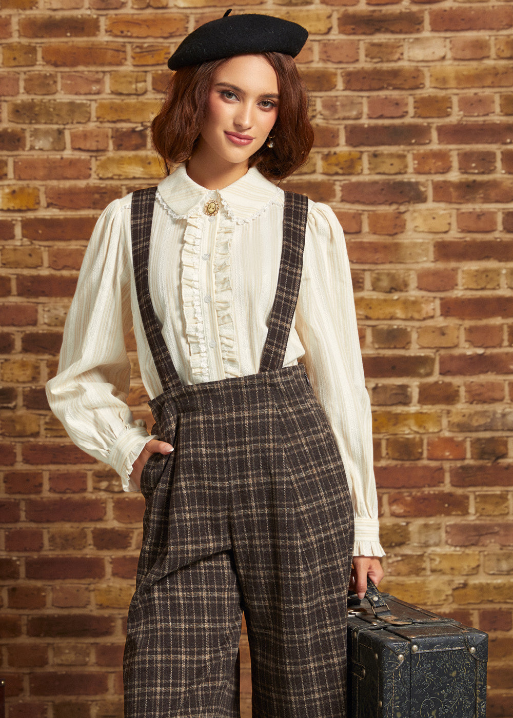 A woman stands against a brick wall wearing a cream ruffled blouse, dark brown plaid suspenders, a black beret, and holding a vintage suitcase.