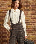 A woman stands against a brick wall wearing a cream ruffled blouse, dark brown plaid suspenders, a black beret, and holding a vintage suitcase.
