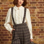 A woman stands against a brick wall wearing a cream ruffled blouse, dark brown plaid suspenders, a black beret, and holding a vintage suitcase.