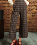 A person wearing brown and beige plaid cropped wide-leg trousers with a white blouse and red Mary Jane heels, standing against a brick wall.
