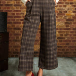 A person wearing brown and beige plaid cropped wide-leg trousers with a white blouse and red Mary Jane heels, standing against a brick wall.