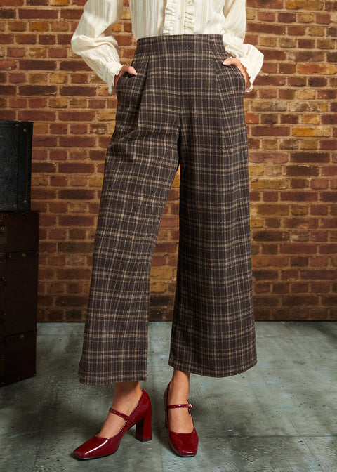 A person wearing brown and beige plaid cropped wide-leg trousers with a white blouse and red Mary Jane heels, standing against a brick wall.