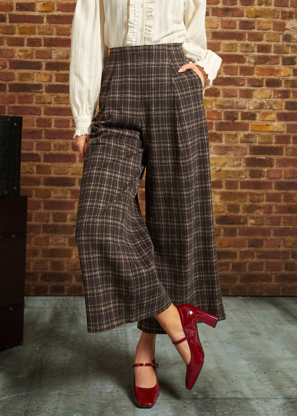 A person wearing brown and beige plaid wide-leg cropped pants with a cream blouse and red Mary Jane heels, standing against a brick wall.