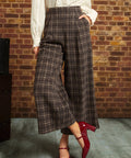 A person wearing brown and beige plaid wide-leg cropped pants with a cream blouse and red Mary Jane heels, standing against a brick wall.