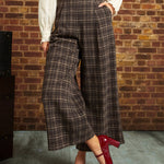 A person wearing brown and beige plaid wide-leg cropped pants with a cream blouse and red Mary Jane heels, standing against a brick wall.