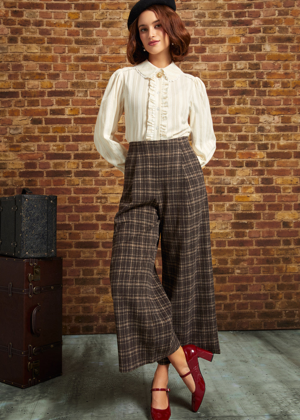 A woman stands in front of a brick wall wearing a cream ruffled blouse, brown plaid wide-leg cropped pants, a black beret, and red heels. A vintage trunk sits to the left.