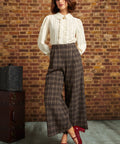 A woman stands in front of a brick wall wearing a cream ruffled blouse, brown plaid wide-leg cropped pants, a black beret, and red heels. A vintage trunk sits to the left.