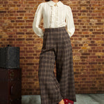 A woman stands in front of a brick wall wearing a cream ruffled blouse, brown plaid wide-leg cropped pants, a black beret, and red heels. A vintage trunk sits to the left.