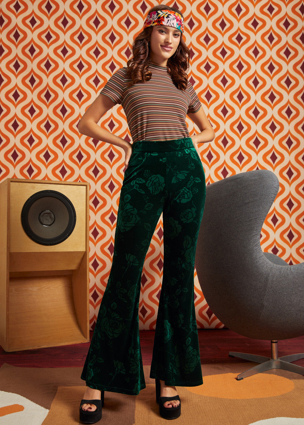 A woman modeling a retro-style outfit featuring dark green velvet flare pants with a tonal floral pattern, paired with a brown and beige striped short-sleeve top and a colorful patterned headband. She is posed in front of a retro orange and brown patterned wall, with a wooden speaker on the left and a gray chair on the right.