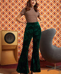A woman modeling a retro-style outfit featuring dark green velvet flare pants with a tonal floral pattern, paired with a brown and beige striped short-sleeve top and a colorful patterned headband. She is posed in front of a retro orange and brown patterned wall, with a wooden speaker on the left and a gray chair on the right.