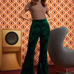 A woman modeling a retro-style outfit featuring dark green velvet flare pants with a tonal floral pattern, paired with a brown and beige striped short-sleeve top and a colorful patterned headband. She is posed in front of a retro orange and brown patterned wall, with a wooden speaker on the left and a gray chair on the right.