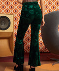 A woman shown from the back wearing dark green velvet flare pants with an allover tonal floral pattern, paired with black platform sandals in a retro-styled room with patterned wallpaper, a speaker, and a chair.