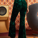 A woman shown from the back wearing dark green velvet flare pants with an allover tonal floral pattern, paired with black platform sandals in a retro-styled room with patterned wallpaper, a speaker, and a chair.