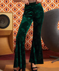 A person wearing dark green velvet flare pants with a tonal floral pattern, paired with black platform sandals, standing in front of a retro orange and white patterned wall.