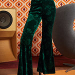 A person wearing dark green velvet flare pants with a tonal floral pattern, paired with black platform sandals, standing in front of a retro orange and white patterned wall.
