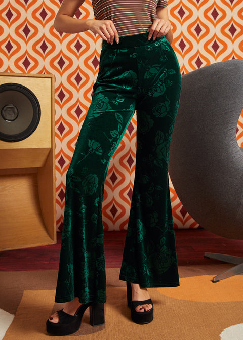 A person wearing dark green velvet flare pants with a tonal floral pattern, paired with black platform sandals, standing in front of a retro orange and white patterned wall.