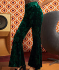 A model wearing ModCloth dark green velvet flare pants with a tonal floral jacquard pattern, paired with black platform sandals, standing in front of a retro orange and white patterned wallpaper background.