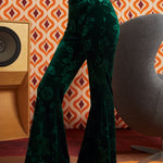 A model wearing ModCloth dark green velvet flare pants with a tonal floral jacquard pattern, paired with black platform sandals, standing in front of a retro orange and white patterned wallpaper background.