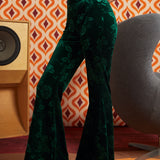 A model wearing ModCloth dark green velvet flare pants with a tonal floral jacquard pattern, paired with black platform sandals, standing in front of a retro orange and white patterned wallpaper background.