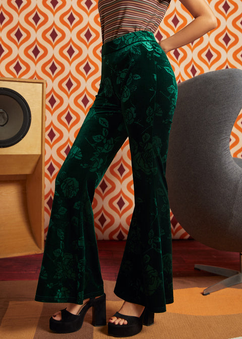 A model wearing ModCloth dark green velvet flare pants with a tonal floral jacquard pattern, paired with black platform sandals, standing in front of a retro orange and white patterned wallpaper background.