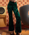A person wearing dark green velvet flare pants with a tonal floral jacquard pattern, paired with black platform heels, standing in front of a retro orange and white patterned wall.