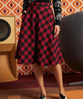 A woman wearing a red and black checkered midi skirt, a black patterned long-sleeve top, and black-and-white loafers, standing in front of a retro orange and white patterned wall with a speaker and a large gray chair.