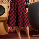 A woman wearing a red and black checkered midi skirt, a black patterned long-sleeve top, and black-and-white loafers, standing in front of a retro orange and white patterned wall with a speaker and a large gray chair.