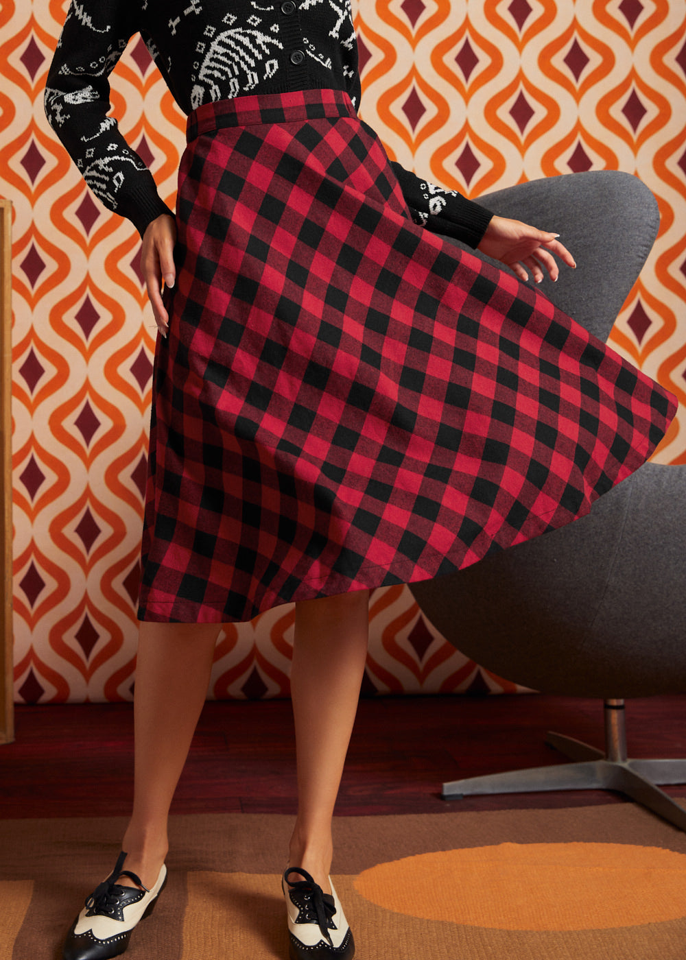 A woman twirling in a ModCloth red and black buffalo plaid A-line skirt, paired with a black and white patterned long-sleeve sweater and black-and-white vintage inspired shoes.