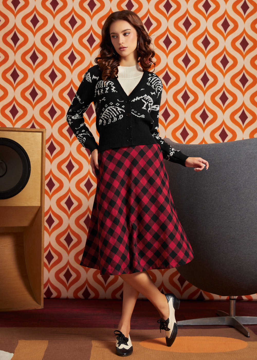 A woman models a black dinosaur bones cardigan and a red and black buffalo plaid midi skirt from ModCloth