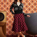 A woman models a black dinosaur bones cardigan and a red and black buffalo plaid midi skirt from ModCloth